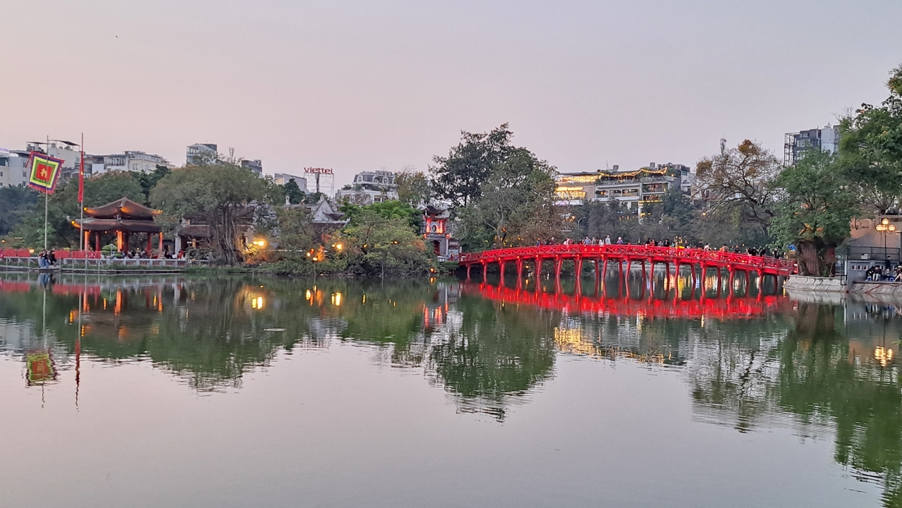 Pont rouge en arc au-dessus d'un étang serein avec un paysage urbain en arrière-plan.