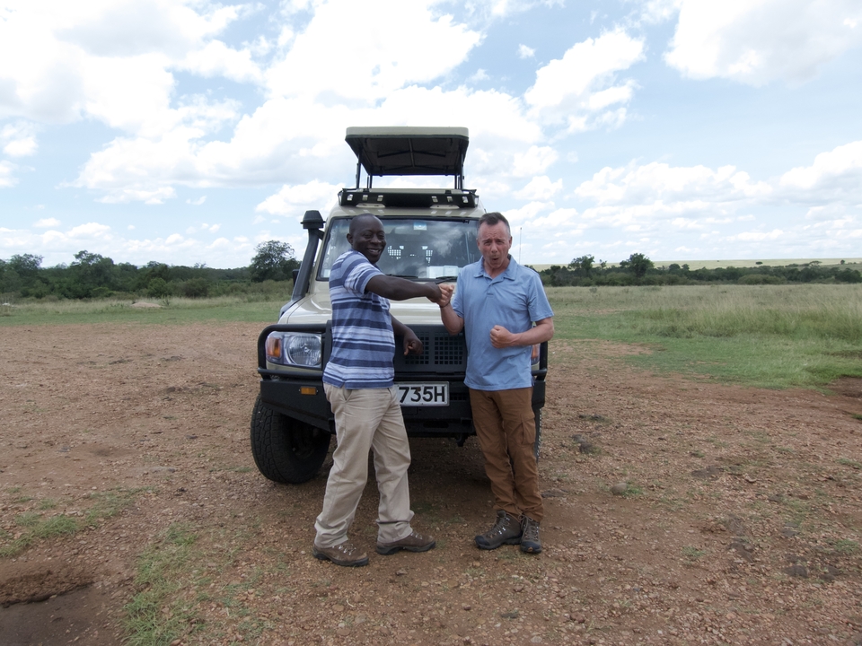 Two men standing in front of a safari vehicle in a savannah setting.