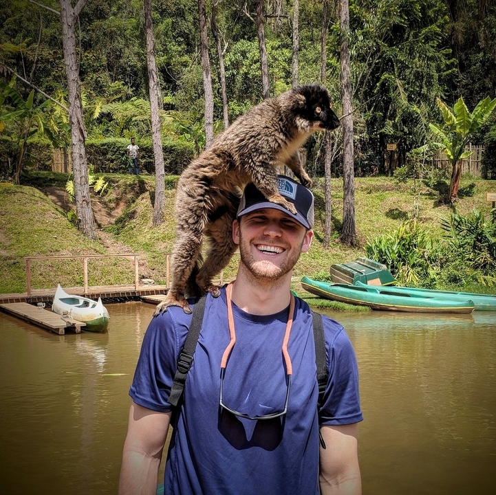 Homme avec un lémurien sur son épaule près d'une rivière avec des canoës.
