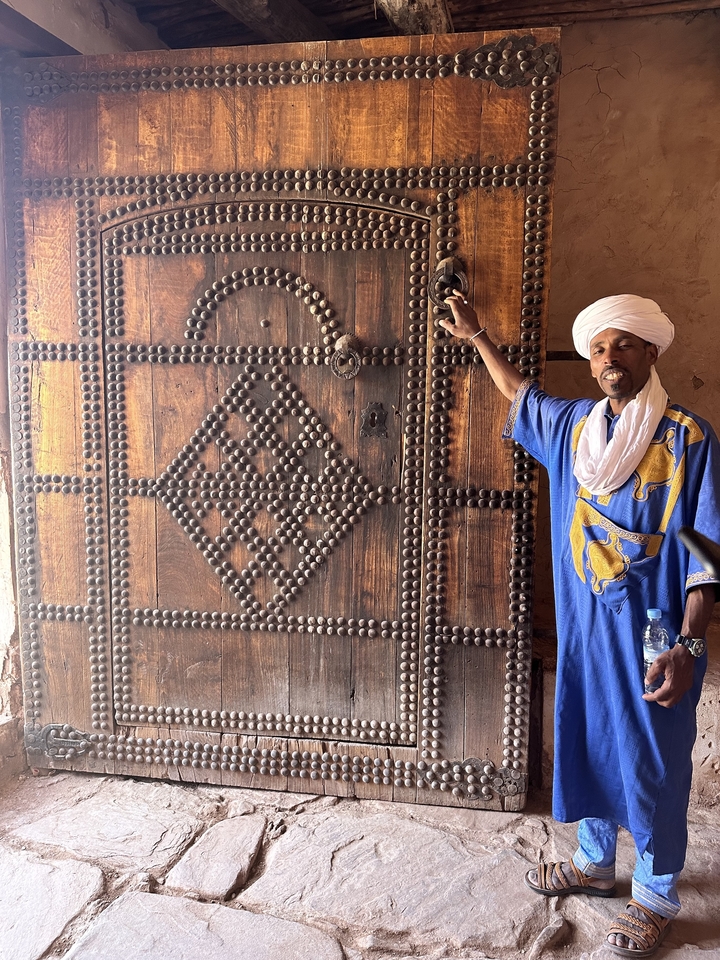 Homme en tenue traditionnelle à côté d'une porte en bois décorée.