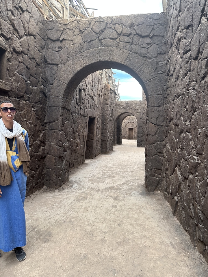 Homme debout dans une ruelle de pierre avec des arches, portant des vêtements traditionnels.