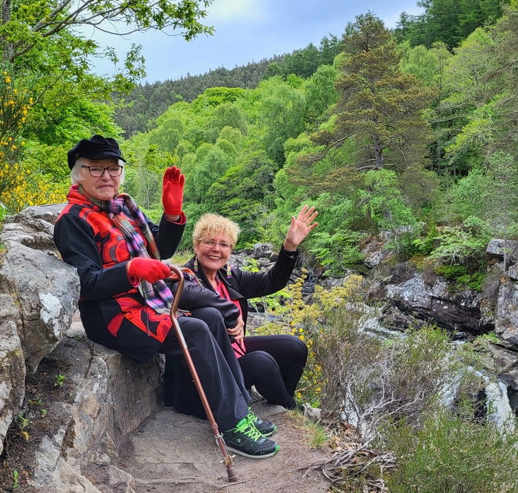 Two women waving amidst a forest landscape.