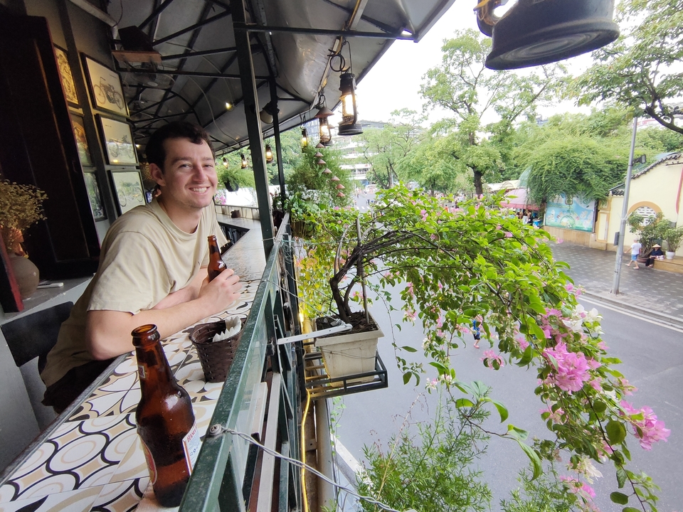 Personne assise sur le balcon d'un café donnant sur une rue avec de la verdure.