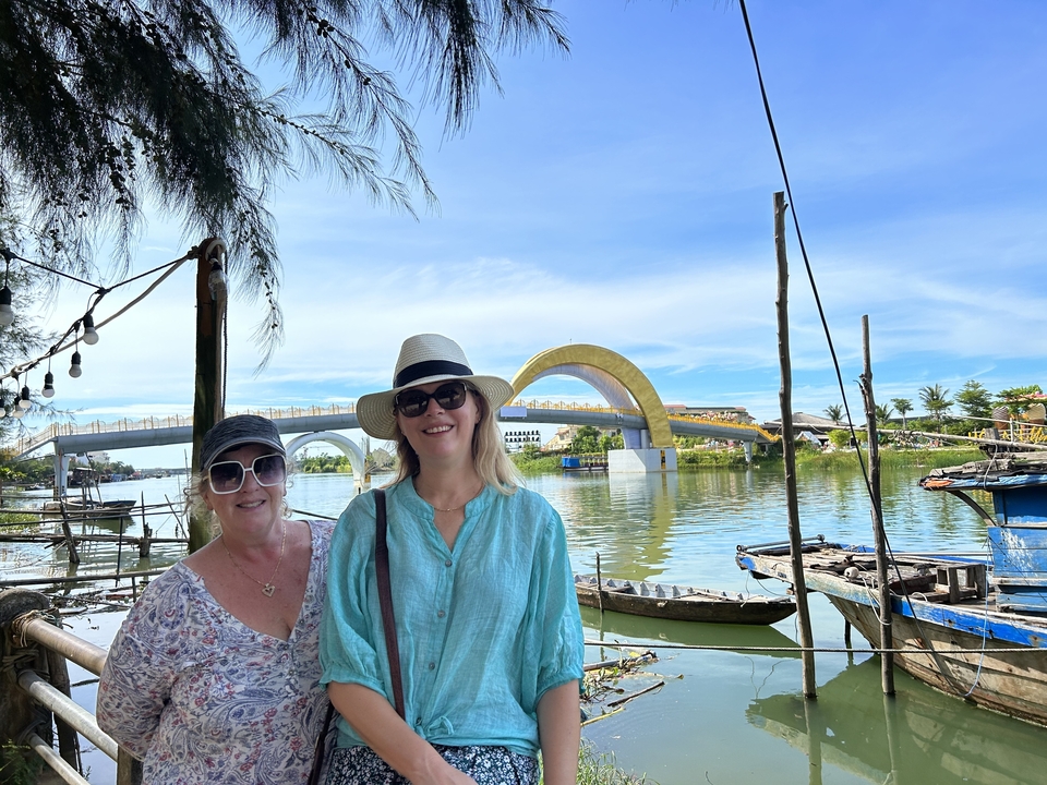Deux femmes debout près d'une rivière avec un pont courbé en arrière-plan.