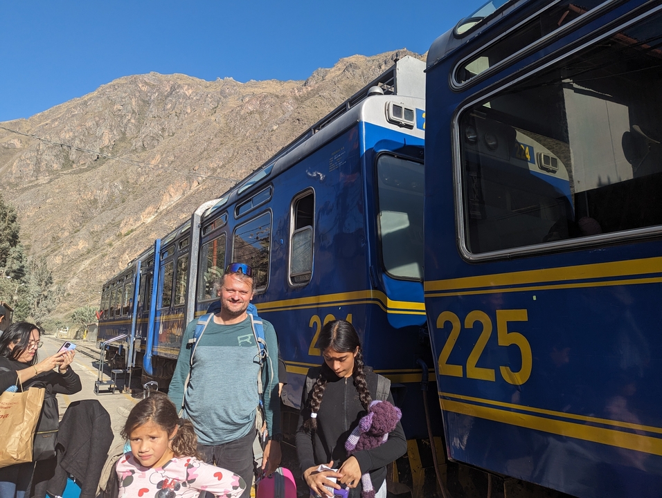 Des gens debout à côté d'un train bleu avec des montagnes en arrière-plan.