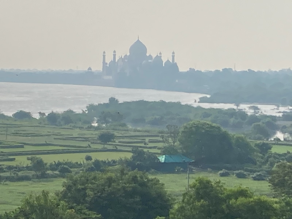 Vue lointaine du Taj Mahal au-dessus d'une rivière avec de la verdure au premier plan.