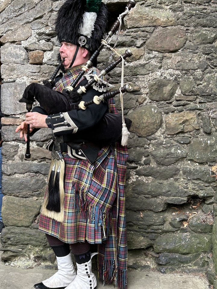 Joueur de cornemuse jouant près d'un mur de pierre en tenue traditionnelle.