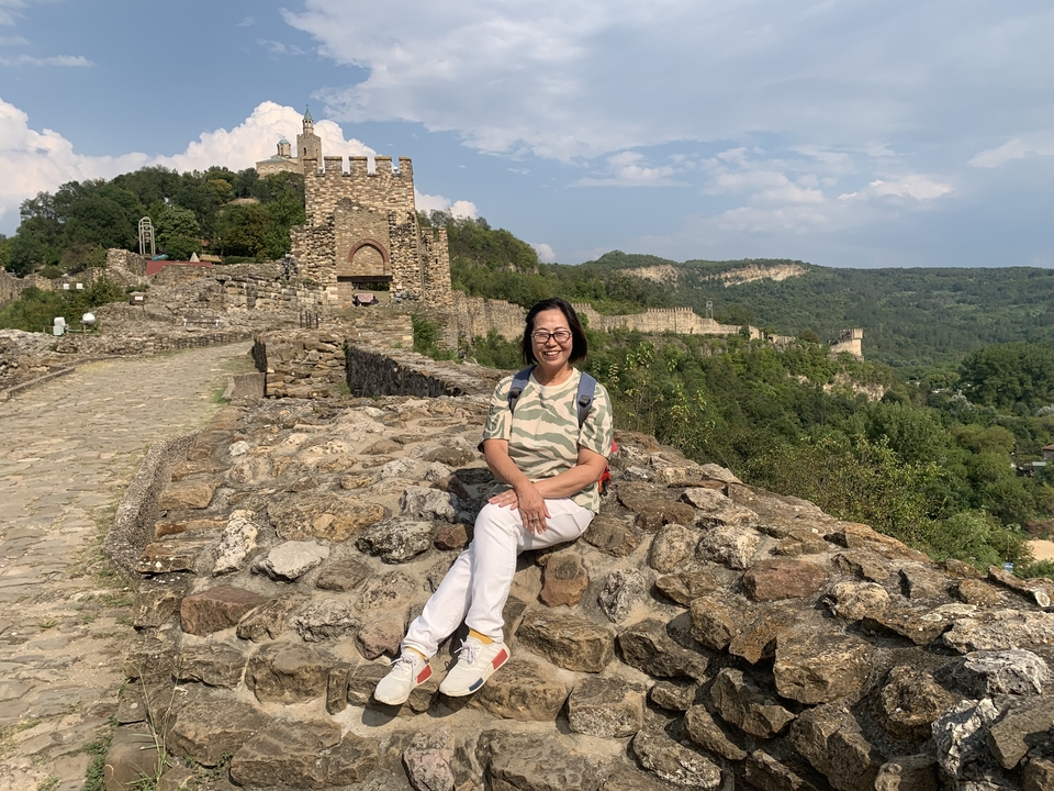 Femme assise sur des ruines de pierre avec une forteresse médiévale en arrière-plan.