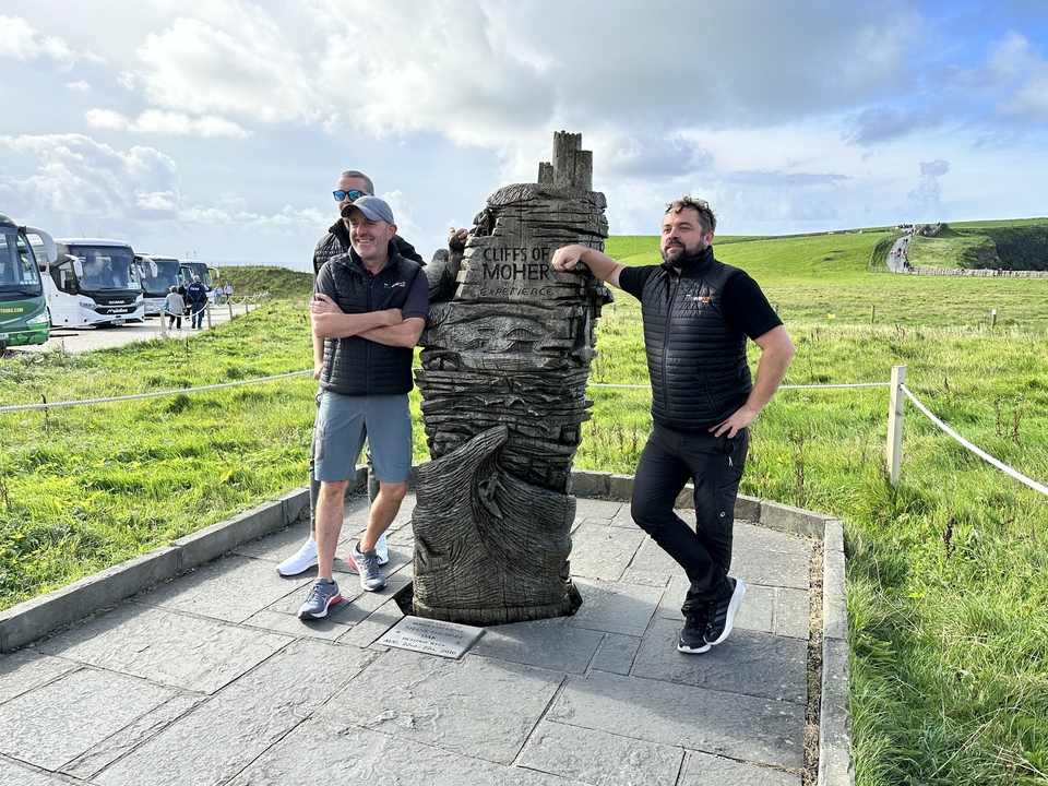 Men posing by a wooden sculpture with scenic cliffs in the background.