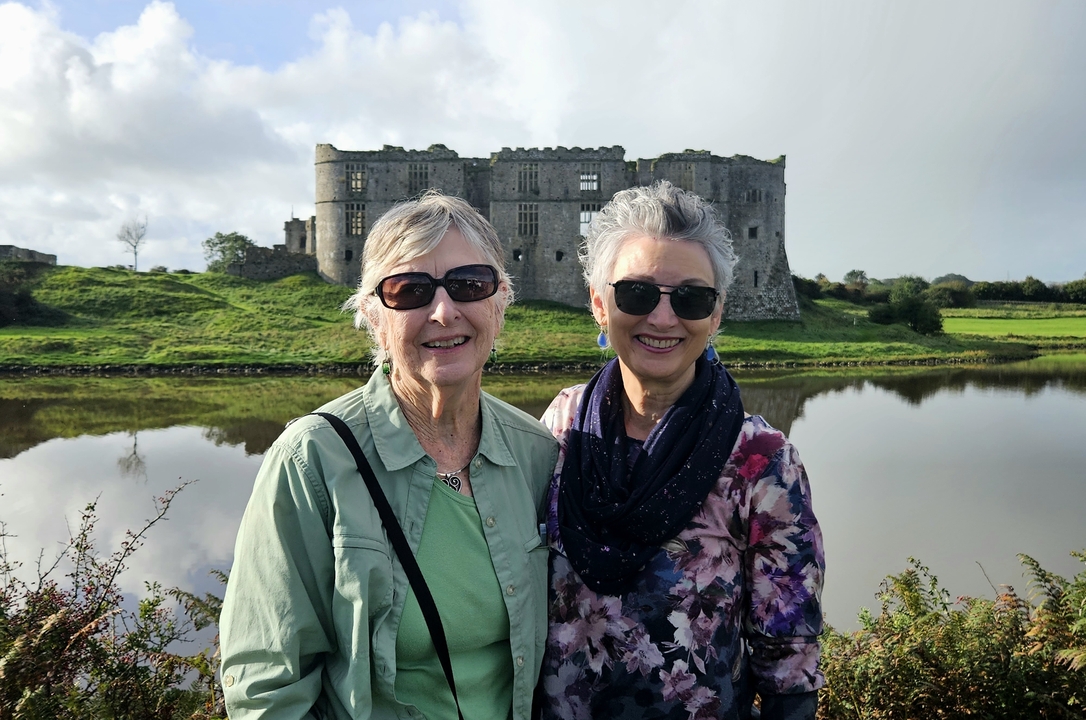 Deux femmes souriantes devant une ruine historique et un lac.