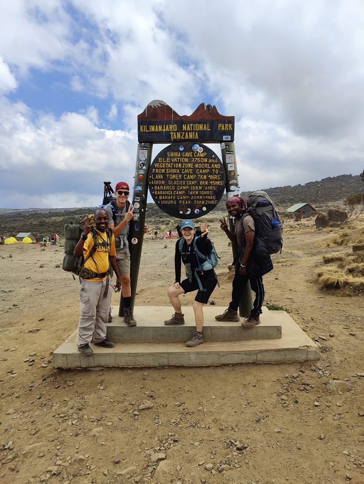 Un groupe de randonneurs souriants au camp de la grotte Shira avec un panneau indicateur.
