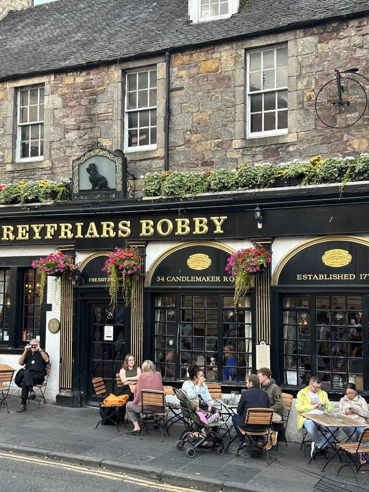 Des gens assis à l'extérieur d'un pub nommé « Greyfriars Bobby » avec des fleurs et un bâtiment en pierre.