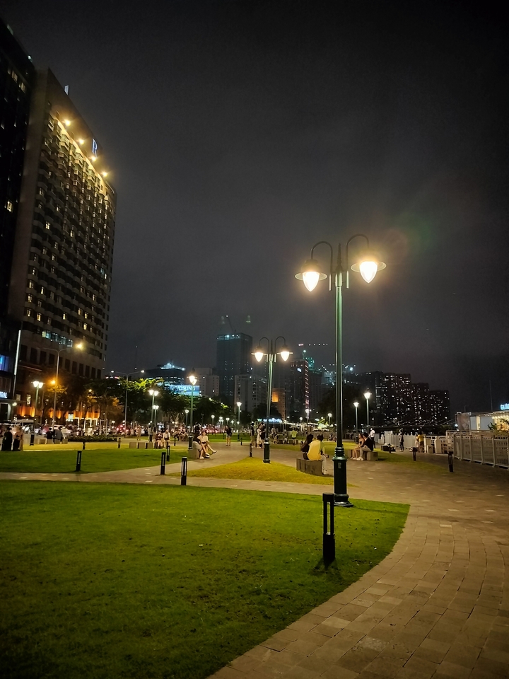 Vue nocturne d'un parc urbain avec des lampadaires éclairés et des immeubles de grande hauteur.