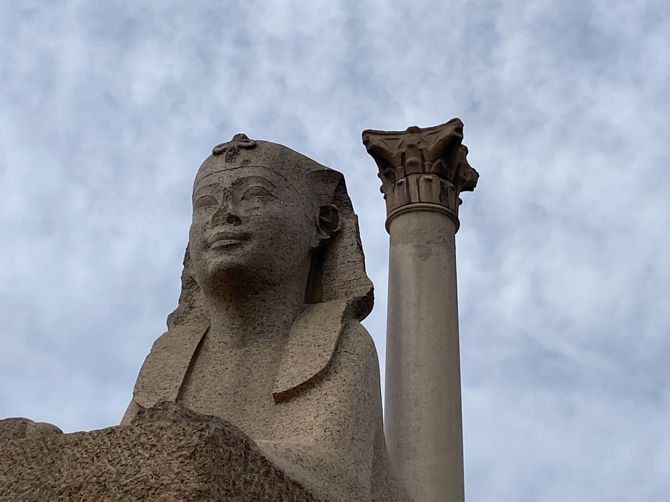 Une statue antique d'un sphinx avec une colonne contre un ciel nuageux.