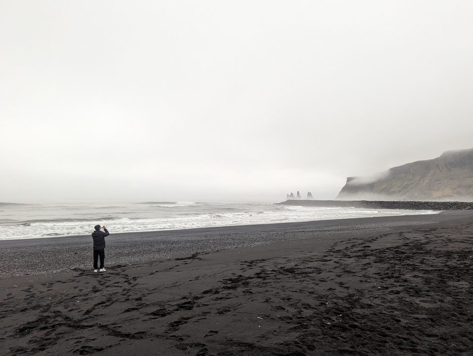 Vue brumeuse d'une plage de sable noir avec une personne photographiant la mer.