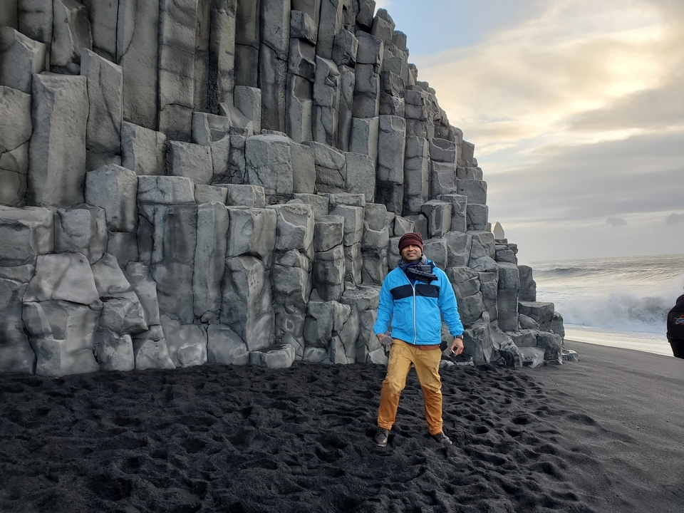 Person standing next to columnar basalt formations on a black beach.