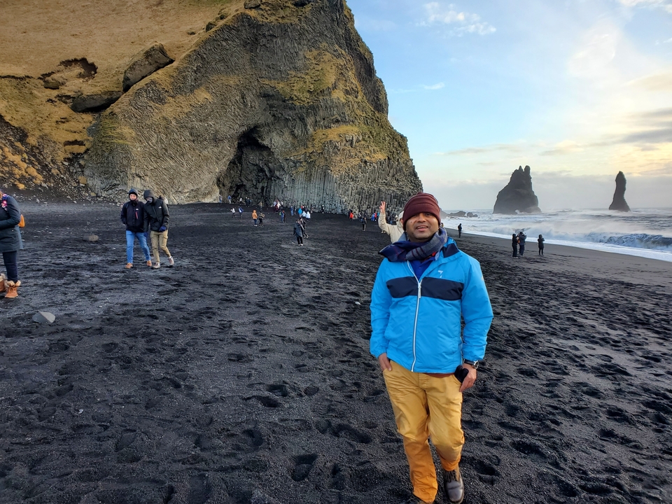 People walking on a black sand beach with dramatic cliffs.