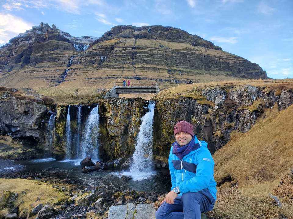 Smiling person in front of scenic waterfalls and mountains.