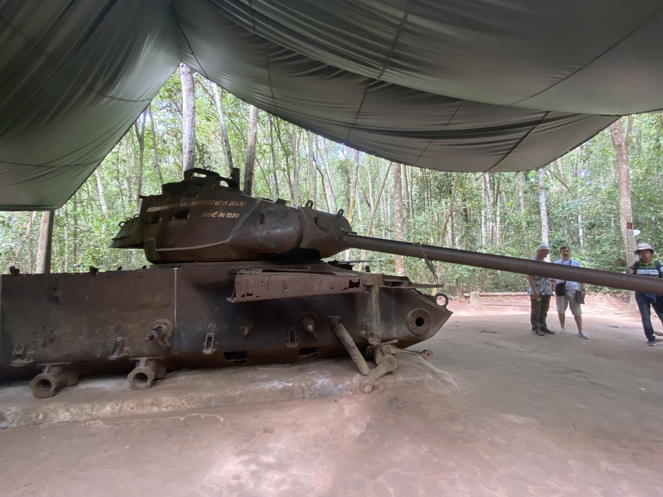 Ancien char militaire exposé sous un auvent avec des visiteurs.
