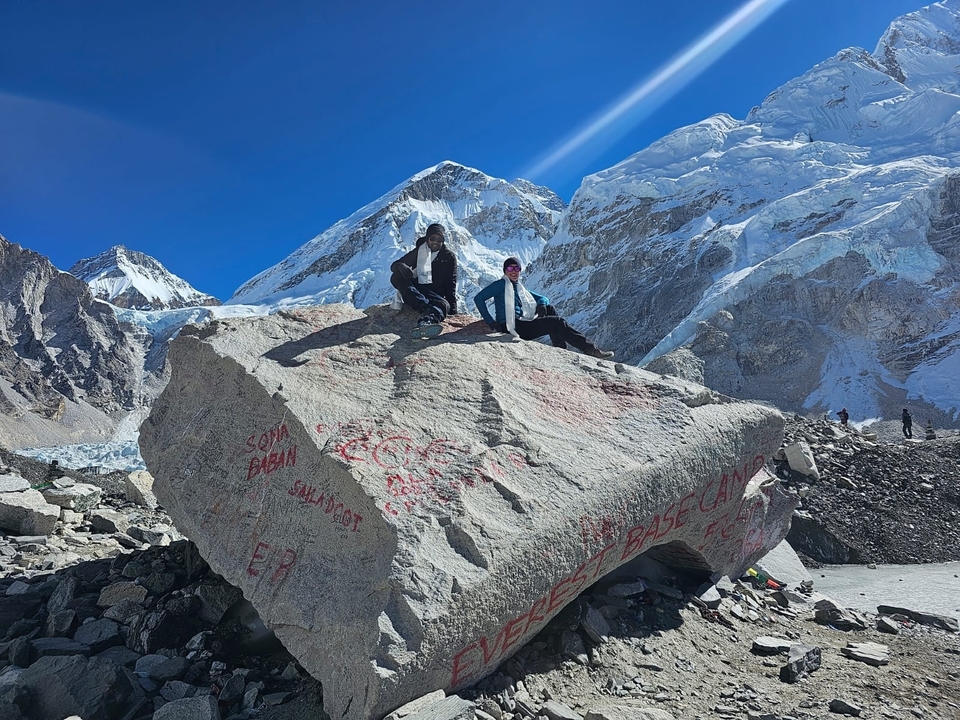 Deux personnes assises sur un gros rocher avec des sommets de montagnes enneigés en arrière-plan.