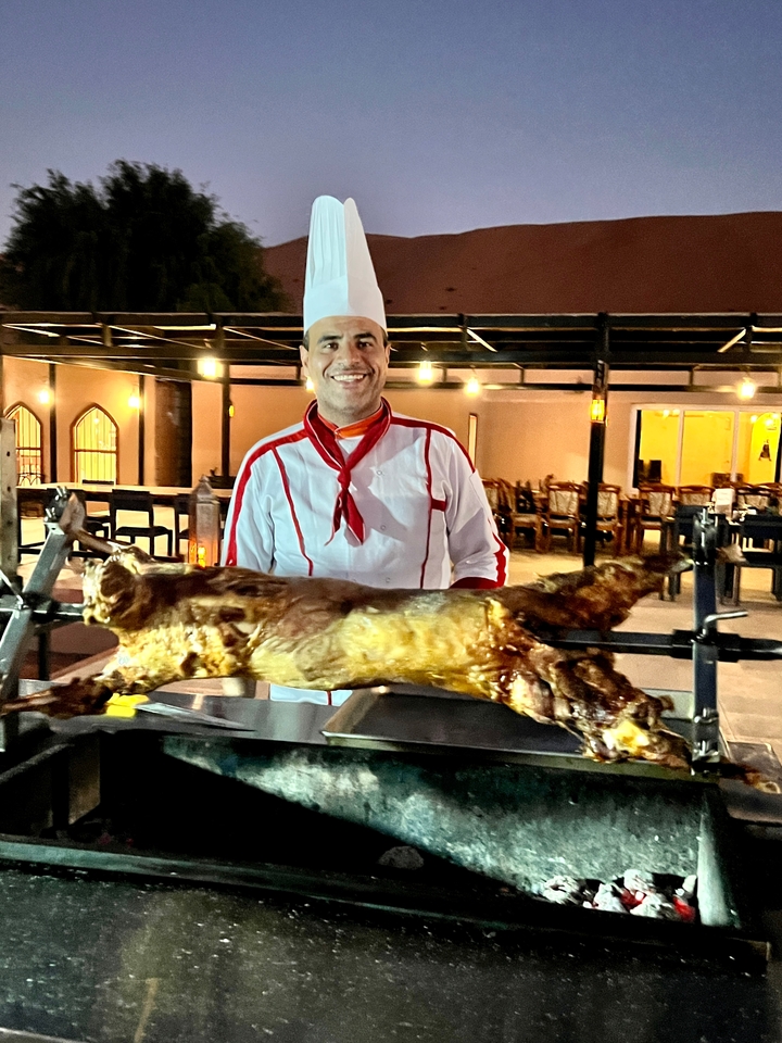 Chef présentant un animal entier rôti à la broche, éclairé par une lumière chaude.