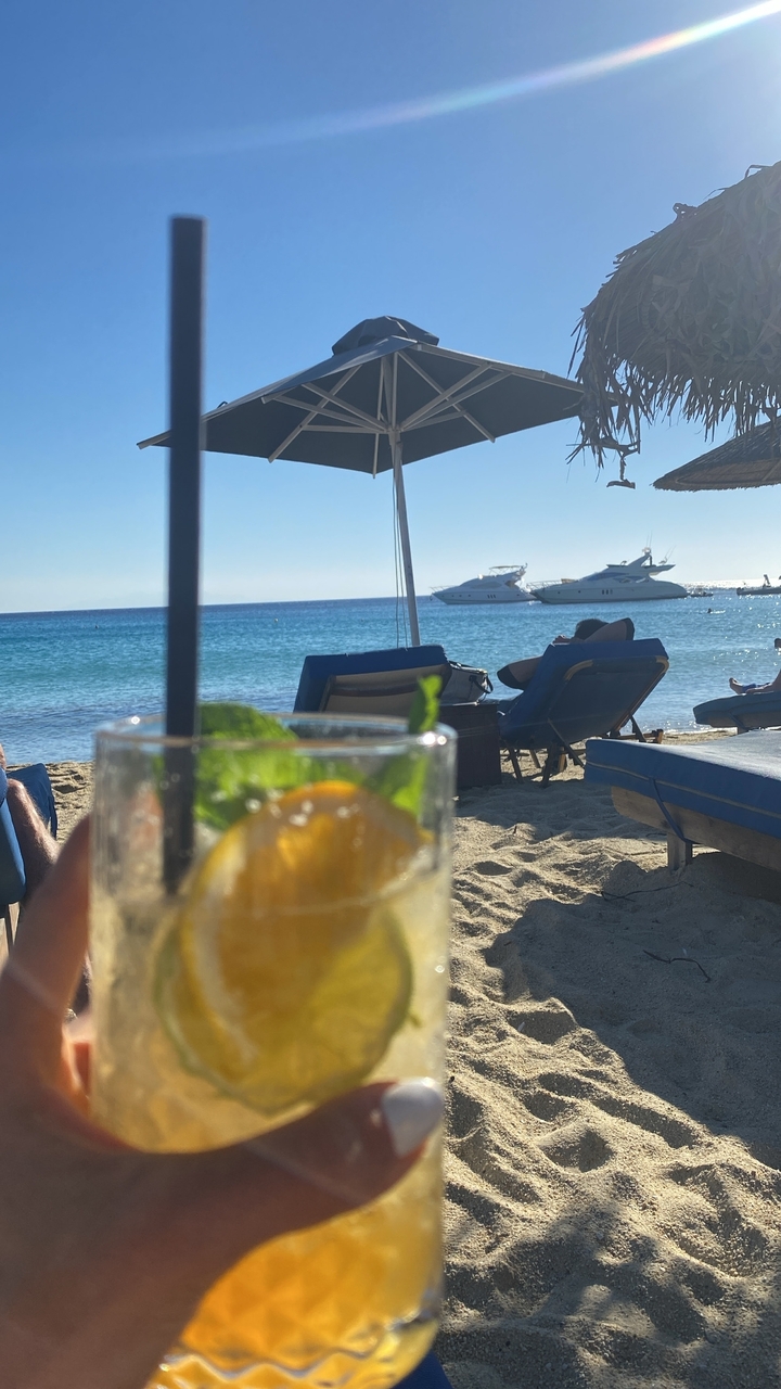 Scène de plage avec transats, parasols et yachts sur l'eau.