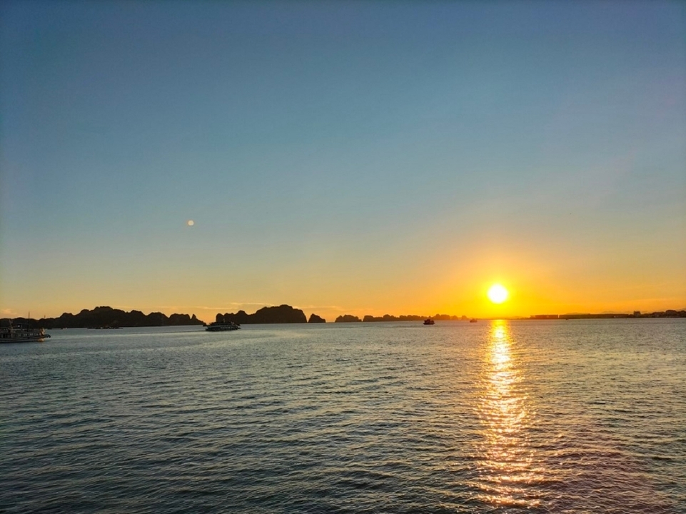 Wide expanse of calm sea with boats in the distance as the sun sets over Halong Bay.