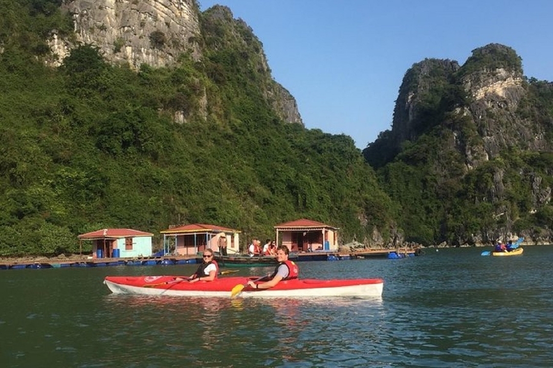 Tourists paddle red kayaks near floating houses against towering limestone cliffs in Halong Bay.