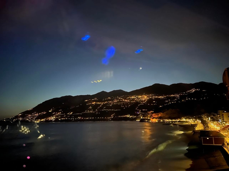 Vue nocturne d'une ville côtière avec des collines illuminées et un croissant de lune.
