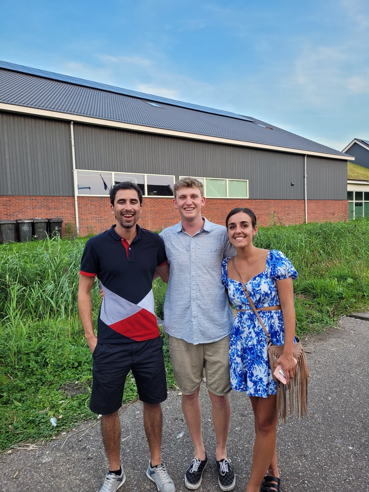 Trois personnes posant devant un champ herbeux et un bâtiment.