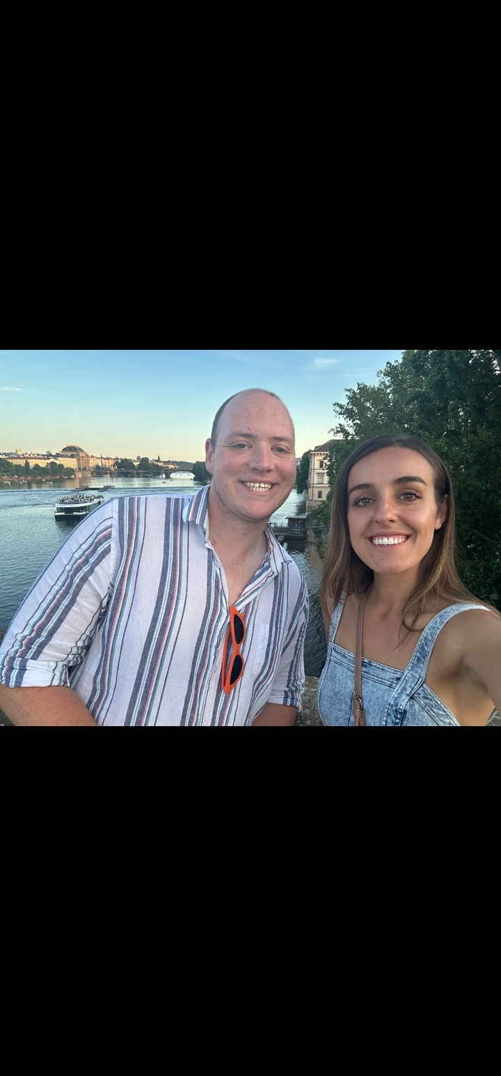 Deux personnes posant près d'une rivière avec un pont et des bâtiments en arrière-plan.