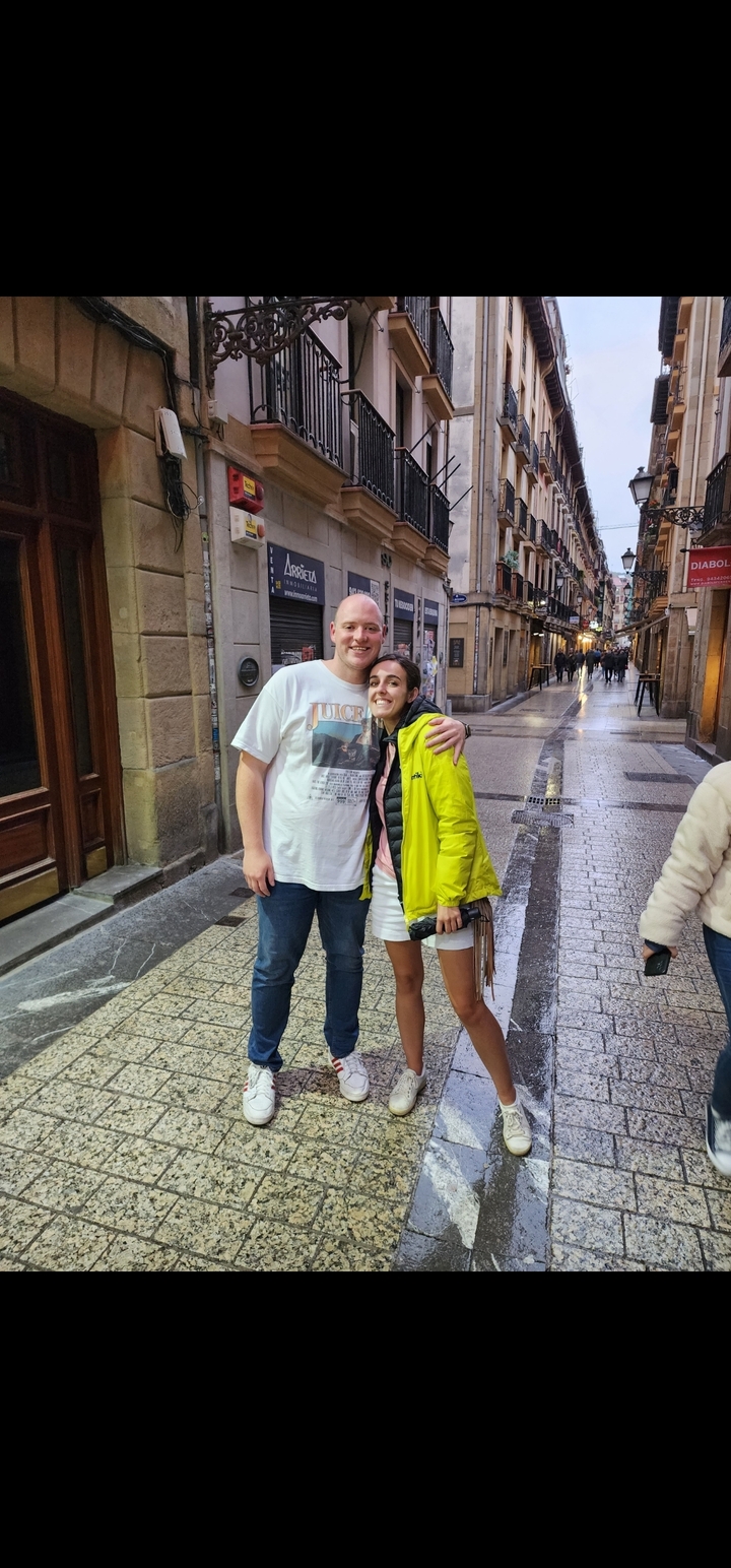 Un couple debout sur une rue pavée avec des bâtiments et des gens en arrière-plan.
