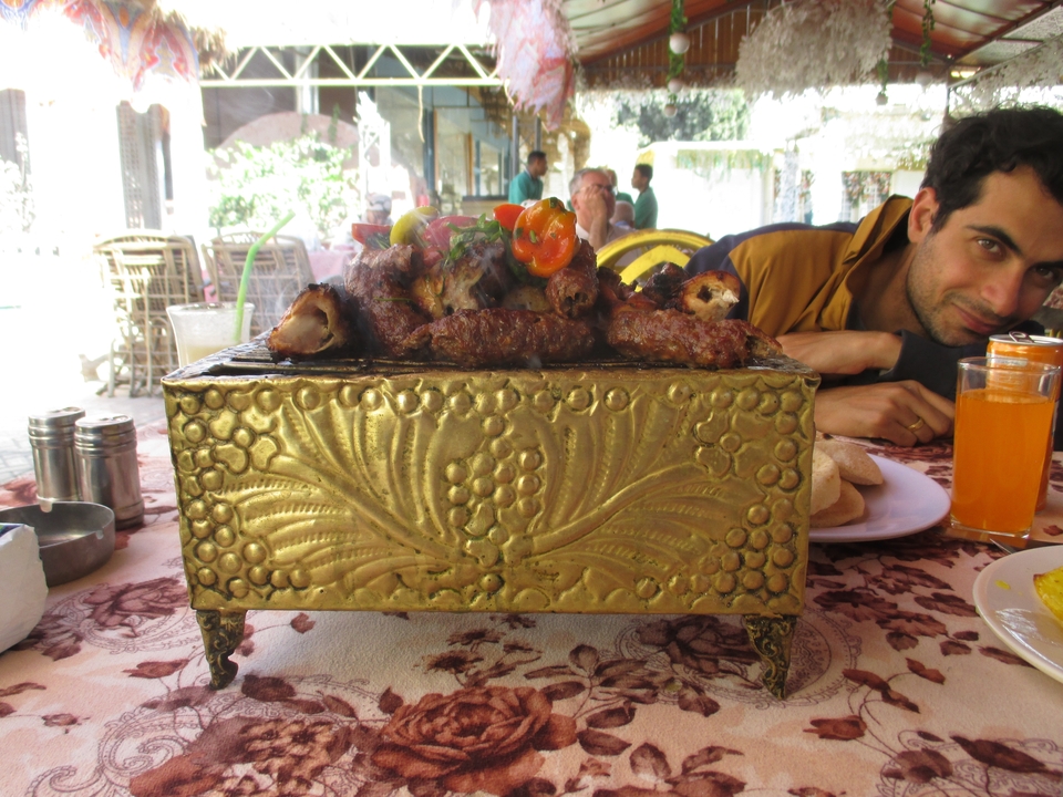 Personne assise à une table avec un récipient métallique décoratif de viande grillée et des boissons.