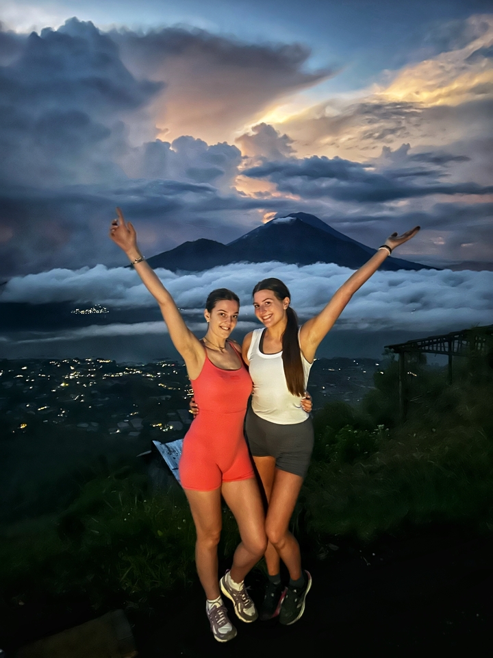 Deux personnes posant avec les bras levés devant un paysage montagneux avec des nuages et les lumières de la ville visibles au loin.
