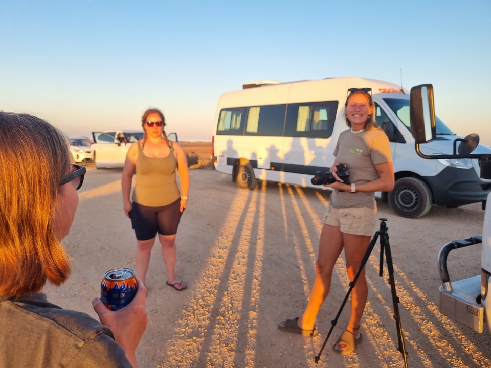 Un groupe de personnes rassemblées près de véhicules blancs sur un chemin de terre avec de longues ombres.