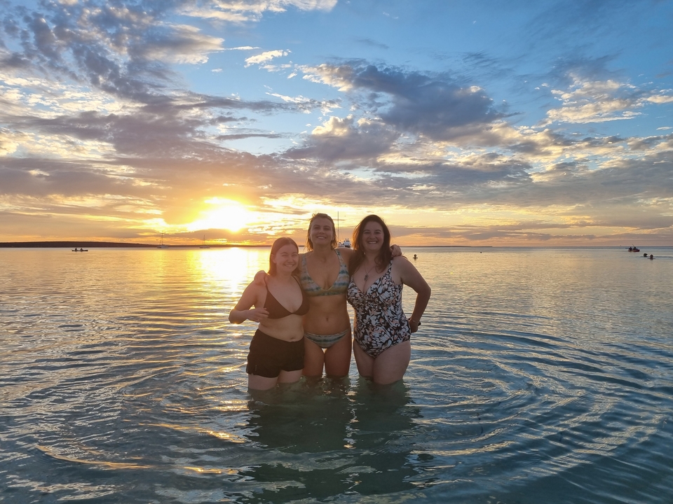 Trois femmes debout dans l'eau peu profonde alors que le soleil se couche derrière elles, créant une scène paisible.