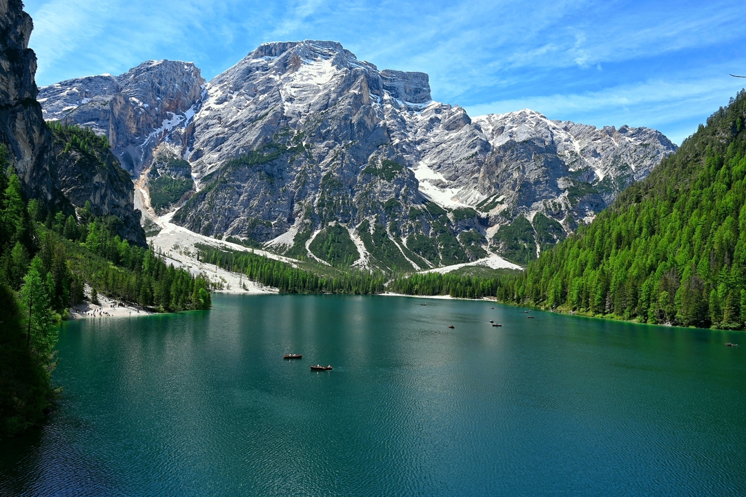 Crystal-clear alpine lake encircled by steep, snow-capped Dolomite peaks and lush green forests under a blue sky.