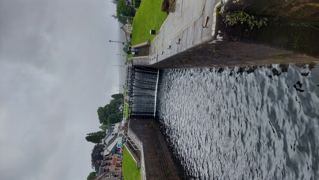 Eau coulant par-dessus une petite écluse ou un barrage avec de la verdure et des bâtiments alentour.