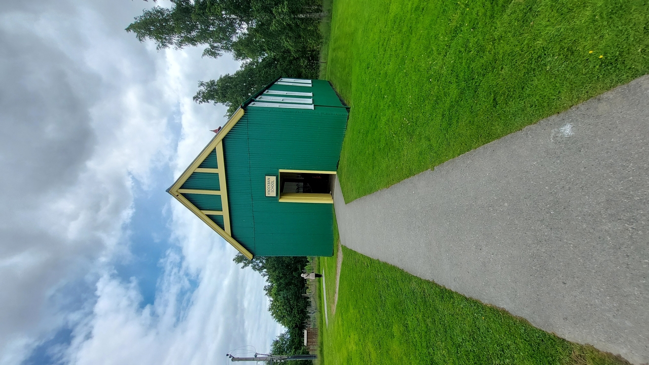Un petit bâtiment vert avec des accents blancs sur un large chemin sous un ciel nuageux.