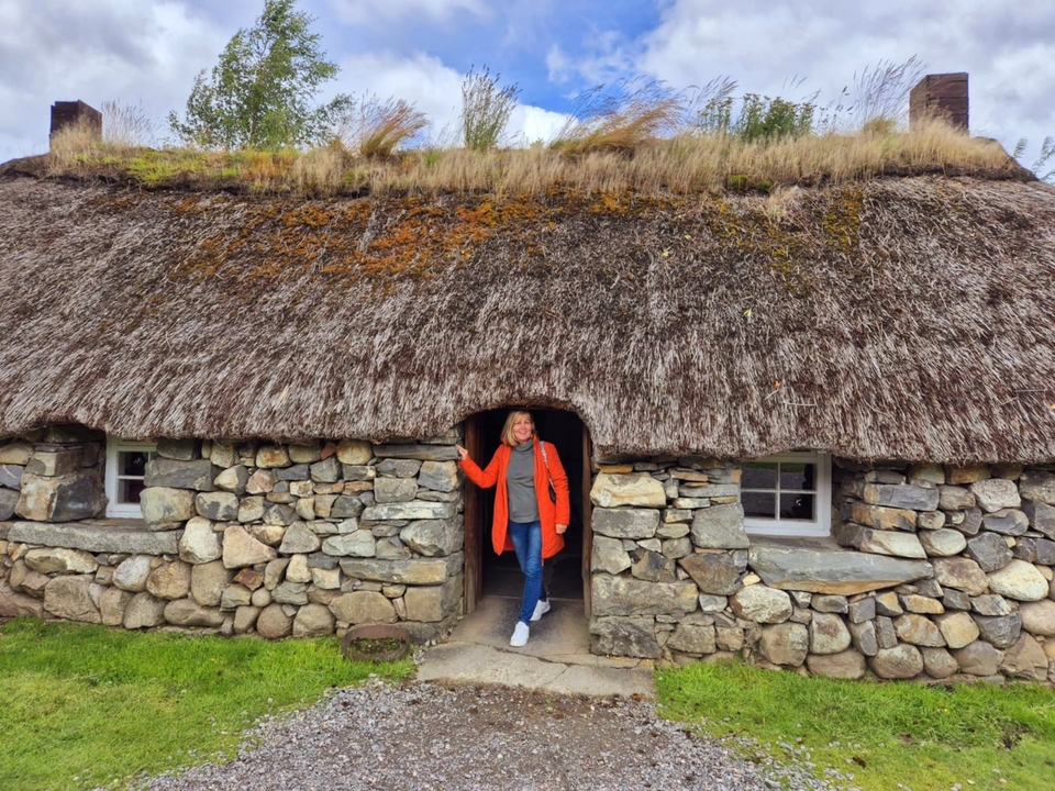 Une personne debout à l'entrée d'une maison traditionnelle en pierre au toit de chaume.