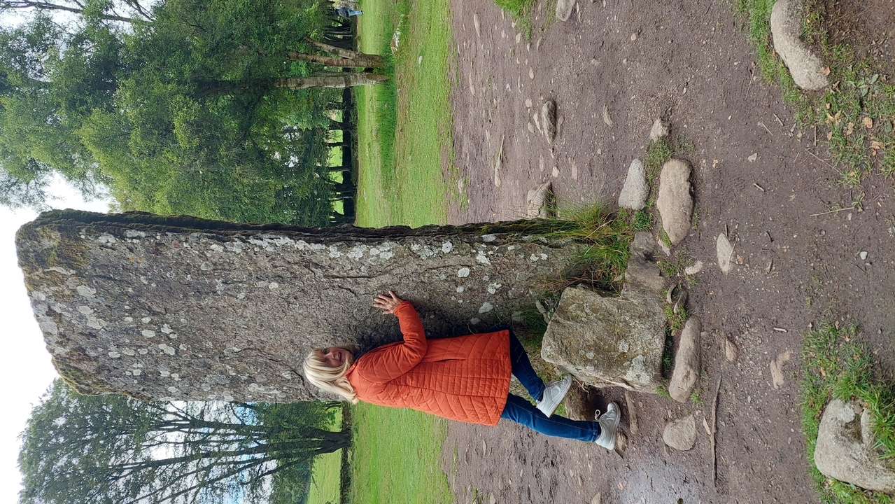 Une personne s'appuyant de manière ludique contre une grande pierre dressée dans une zone herbeuse.