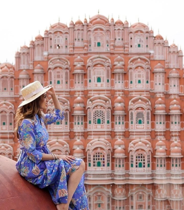 Une femme avec un chapeau pose devant une façade rose avec de nombreuses fenêtres.