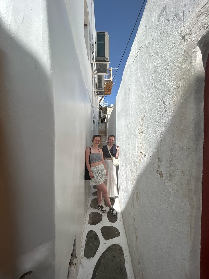 Deux femmes debout dans une ruelle étroite aux murs blancs sous un ciel bleu.