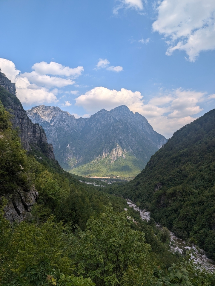 Vallée montagneuse avec des pics et des forêts sous un ciel bleu avec des nuages.