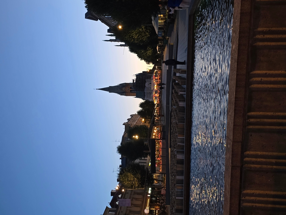 Une vue pittoresque avec une haute flèche et une rivière, beaucoup de lumières suggérant que c'est le soir.