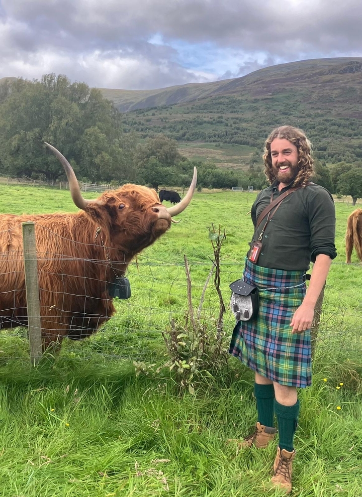 Homme en tenue écossaise traditionnelle avec une vache Highland en arrière-plan.