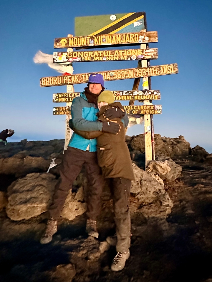 "Couple qui s'enlace au sommet avec un panneau indiquant le pic."
Alternative translation: "Couple s'embrassant au sommet avec un panneau marquant le pic."