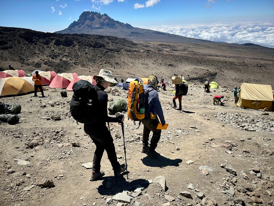 Des randonneurs avec des sacs à dos passent devant des tentes sur un terrain rocheux.
