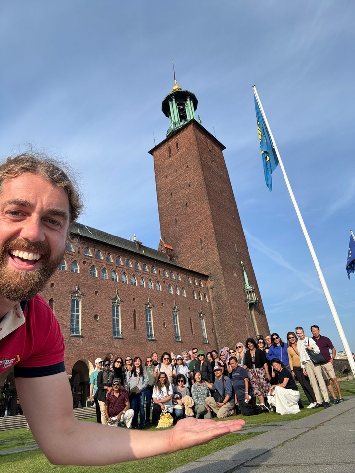 Personne souriante devant un bâtiment en briques rouges avec des drapeaux.