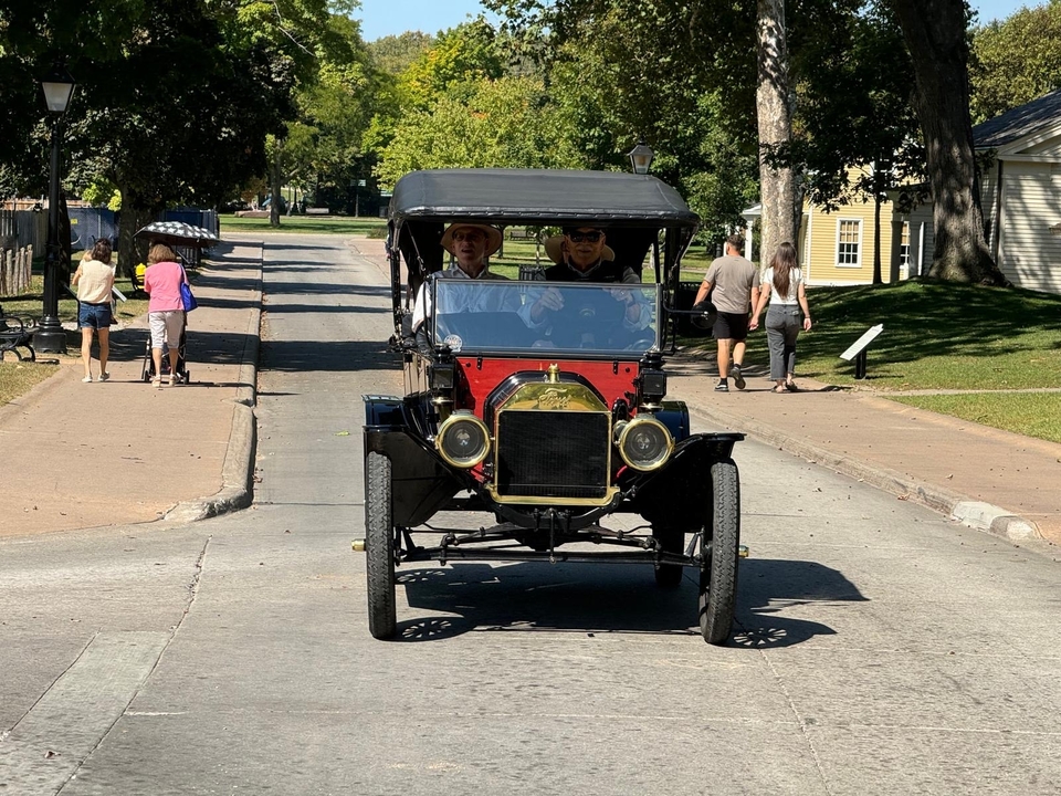 Voiture ancienne roulant dans une rue avec des maisons et des gens qui marchent.